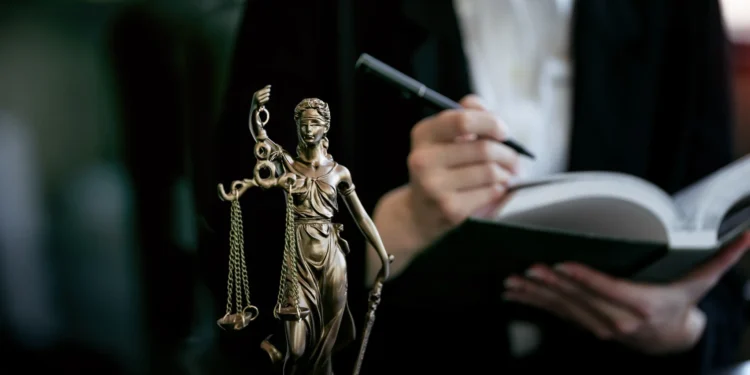 Estátua de Lady Justice com balança, enquanto advogado anota em livro, representando o universo jurídico.