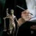Estátua de Lady Justice com balança, enquanto advogado anota em livro, representando o universo jurídico.