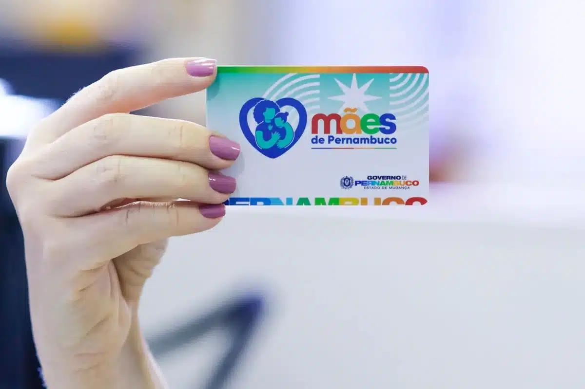 Calendário de março do Mães de Pernambuco confirma pagamento para dia 9. Cartão do programa Mães de Pernambuco exibido em mão feminina com informação sobre pagamento de março de 2026.