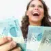 Pessoa sorrindo segurando cédulas de R$ 100,00, relacionada ao pagamento do auxílio de R$ 1.000,00 da Caixa para CPFs com finais de 0 a 9, nos dias 26 e 27 de fevereiro de 2026.