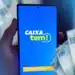 Imagem do aplicativo Caixa Tem sendo acessado em um smartphone, com dinheiro em notas ao fundo. O aplicativo vai liberar R$ 1.200 para diversos brasileiros até 5 de março.