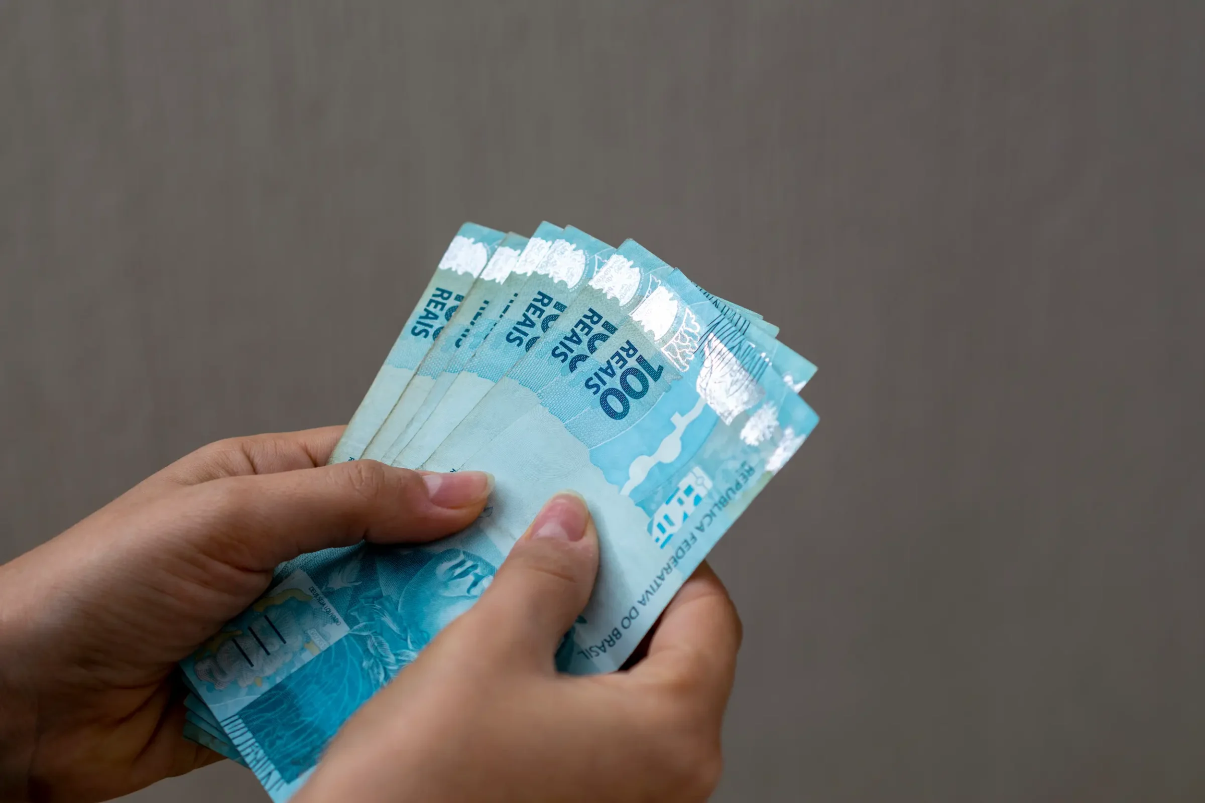 Auxílio financeiro disponibilizado aos estudantes Mãos segurando notas de cem reais representando auxílio financeiro estudantil.