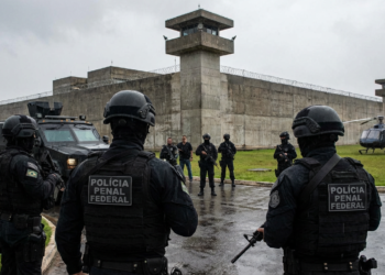 Policiais penais federais armados cercam presídio com helicóptero e veículos blindados ao fundo