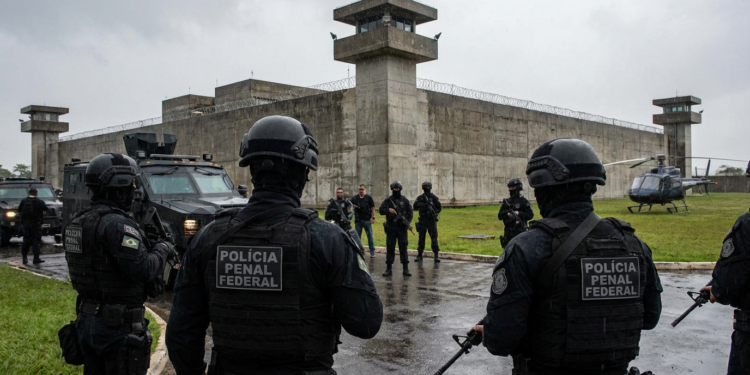Policiais penais federais armados cercam presídio com helicóptero e veículos blindados ao fundo
