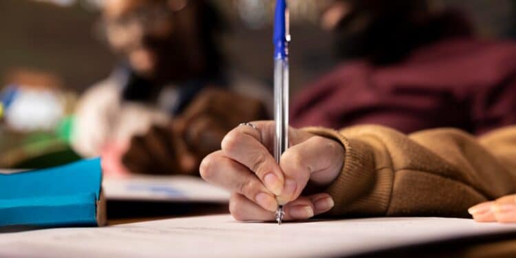 Mão segurando caneta esferográfica azul escrevendo em folha de papel representando candidato realizando prova do processo seletivo de Ubiratã PR