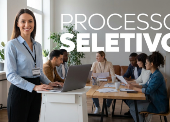 Mulher sorridente em pé ao lado de notebook liderando processo seletivo com equipe reunida ao fundo