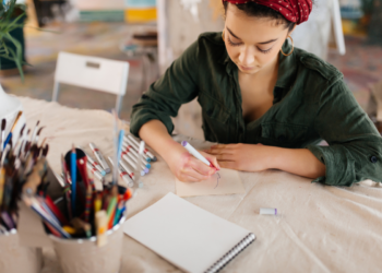 Mulher segurando marcador vermelho desenha em papel sobre mesa cheia de materiais artísticos