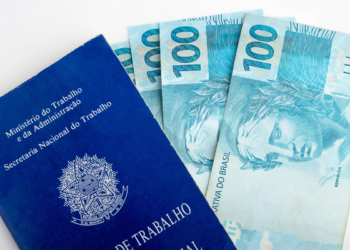 Carteira de trabalho azul posicionada ao lado de três notas de 100 reais sobre superfície branca