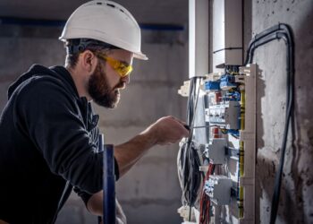 Um eletricista usando capacete branco e óculos de proteção amarelados, focado em conectar fios em um painel elétrico com disjuntores e fiações coloridas.