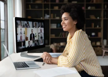 Mulher sorrindo, participando de uma videoconferência no home office, com vários colegas de trabalho na tela do computador.