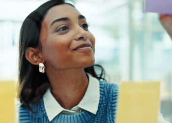 Mulher sorrindo enquanto organiza post-its em um painel de vidro, simbolizando planejamento de carreira.