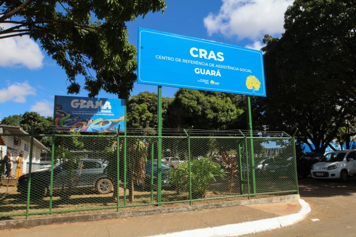 Sedes DF Placa azul do CRAS Guará em destaque sobre cerca, com árvores e carros estacionados ao fundo