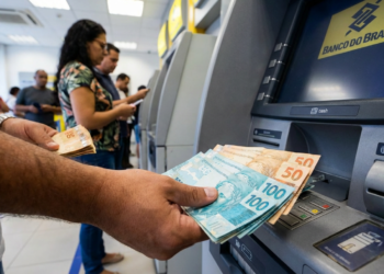 Pessoa segurando notas de R$ 100 e R$ 50 ao retirar dinheiro em caixa eletrônico no banco