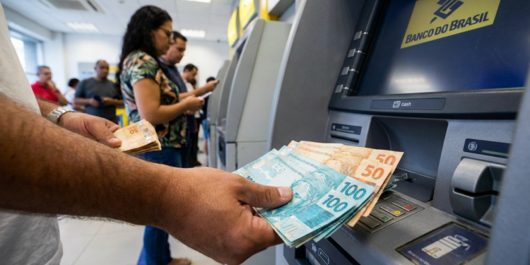 Pessoa segurando notas de R$ 100 e R$ 50 ao retirar dinheiro em caixa eletrônico no banco