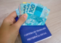 Mão segura notas de reais sobre uma carteira de trabalho azul em superfície de madeira clara