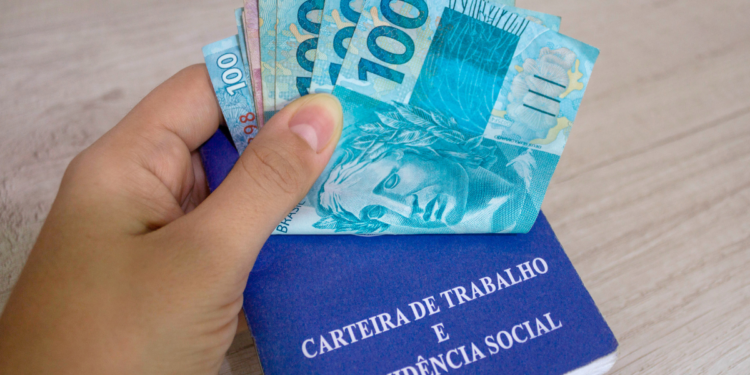 Mão segura notas de reais sobre uma carteira de trabalho azul em superfície de madeira clara