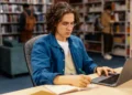 Estudante com laptop e caderno, focado no TCC em uma biblioteca.