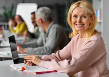 Mulher loira sorridente na faixa dos 50 anos, sentada com um notebook e caderno em um ambiente de escritório compartilhado, representando o sucesso em profissões autônomas.