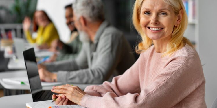 Mulher loira sorridente na faixa dos 50 anos, sentada com um notebook e caderno em um ambiente de escritório compartilhado, representando o sucesso em profissões autônomas.