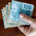 Mão segurando notas de R$ 100 e R$ 50 em leque sobre fundo marrom.
