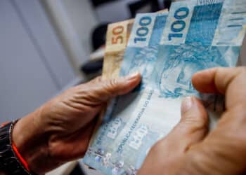 Mãos contando notas de R$ 100 e R$ 50 em ambiente interno