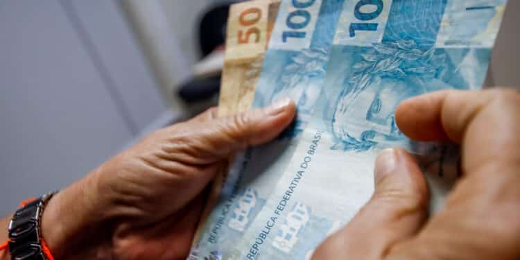 Mãos contando notas de R$ 100 e R$ 50 em ambiente interno
