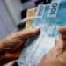 Mãos contando notas de R$ 100 e R$ 50 em ambiente interno