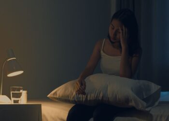Mulher jovem asiática com dor de cabeça à noite, sofrendo de insônia enquanto tenta dormir em um ambiente com pouca luz. Estudo científico revela que dormir tarde pode afetar o coração a longo prazo.