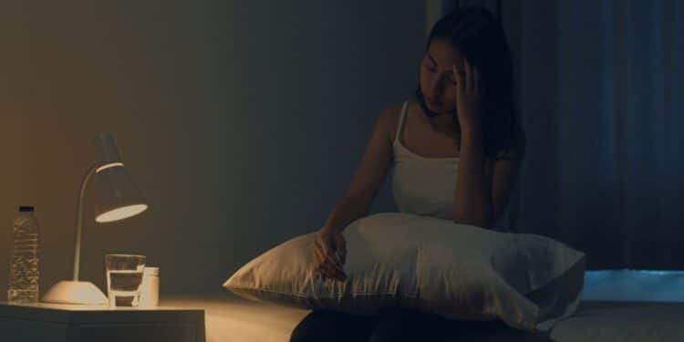 Mulher jovem asiática com dor de cabeça à noite, sofrendo de insônia enquanto tenta dormir em um ambiente com pouca luz. Estudo científico revela que dormir tarde pode afetar o coração a longo prazo.