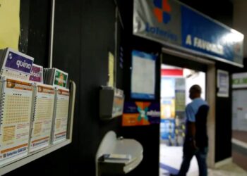 Imagem de uma casa lotérica, com apostadores observando os bilhetes disponíveis para jogos como Quina, Lotomania e outros.
