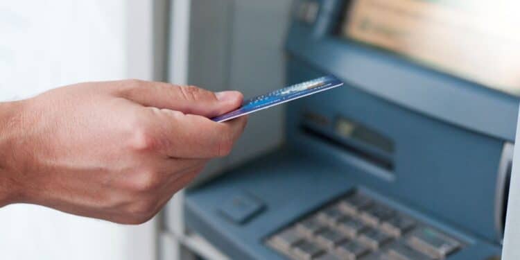 Mão segurando cartão de banco próxima à entrada de caixa eletrônico.