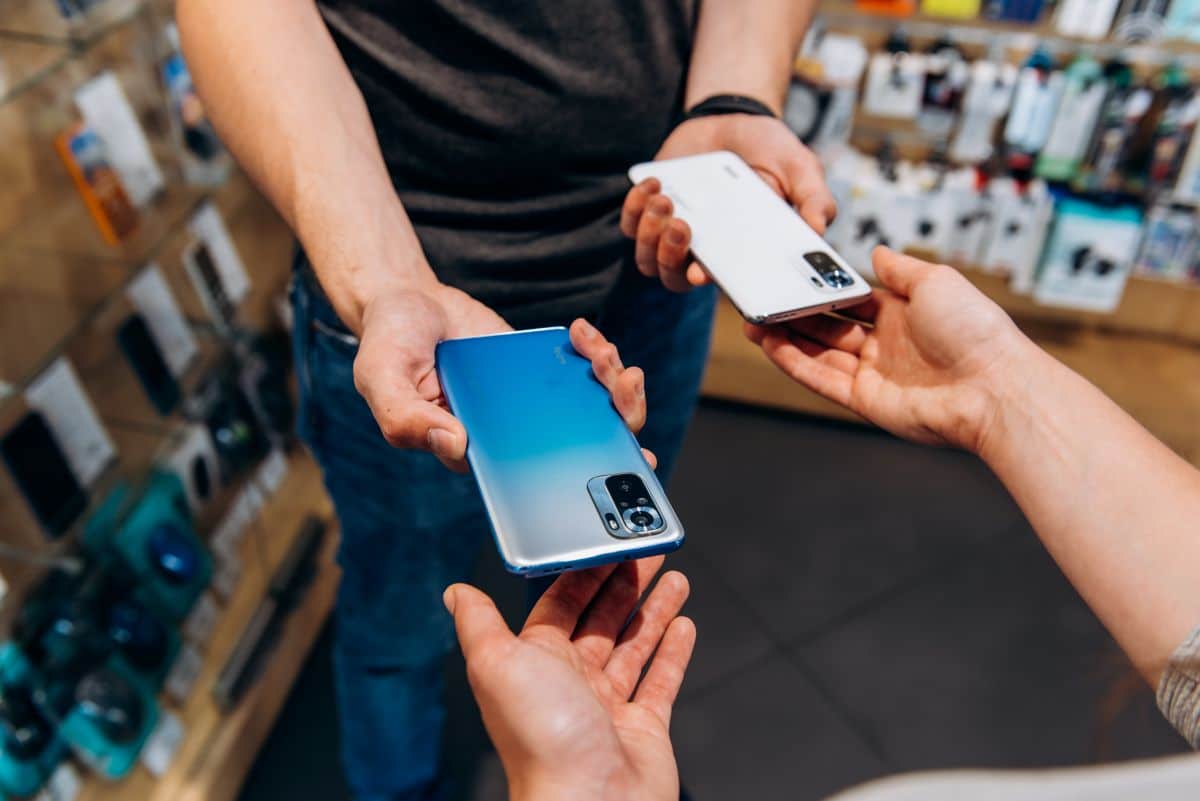 Duas pessoas trocando smartphones em uma loja de eletrônicos, com celulares visíveis em mãos, destacando um modelo em azul e outro em branco.