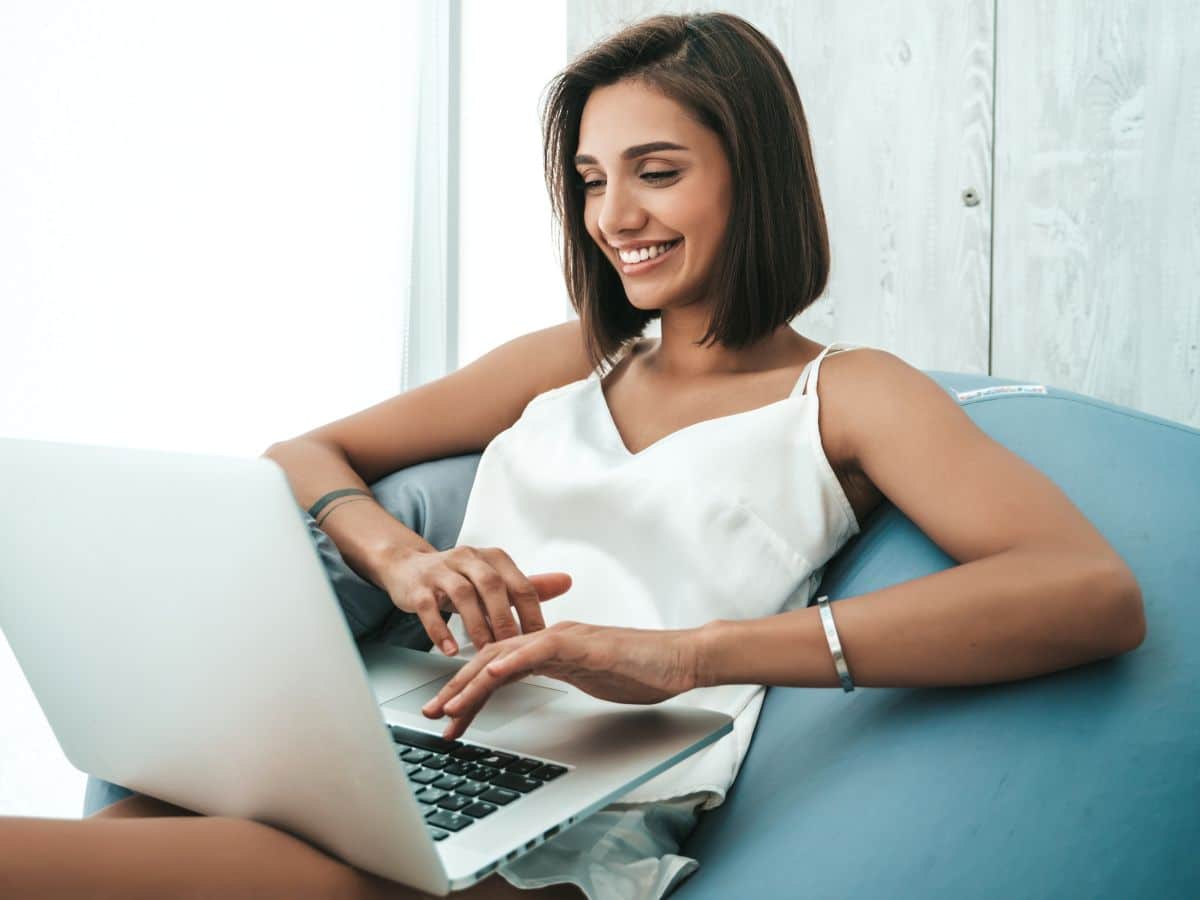 Mulher sorridente sentada em puff azul digitando em notebook prateado.