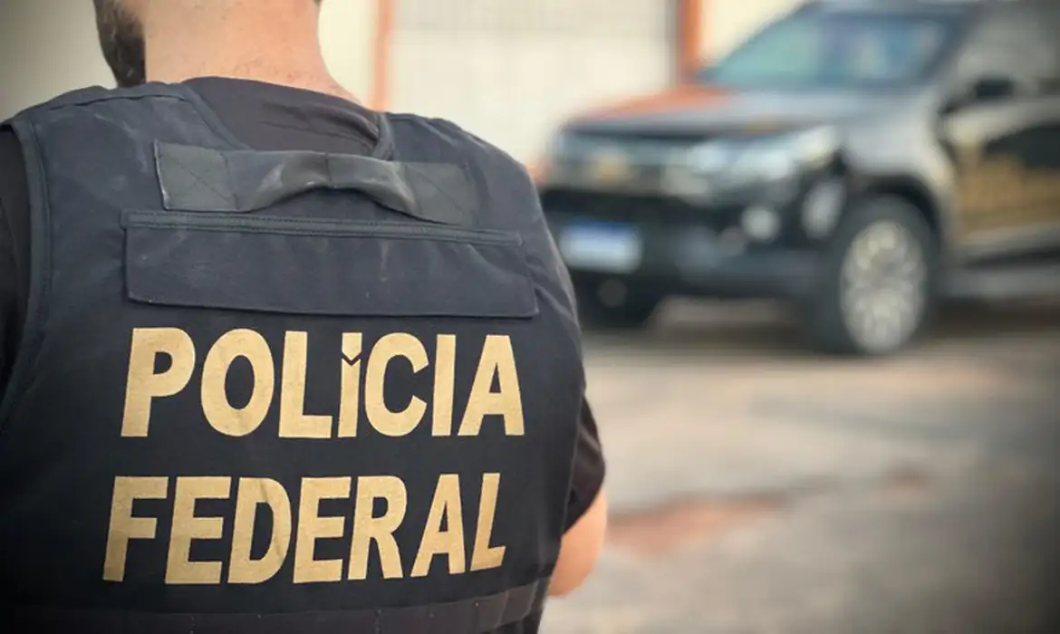Agente da Polícia Federal com colete identificado, em frente a um veículo da corporação.