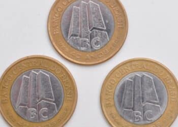 Fotografia de três moedas de 1 Real comemorativas dos 40 anos do Banco Central, organizadas em formato de triângulo sobre um fundo branco. A imagem exibe o lado do anverso das moedas, destacando os detalhes do núcleo prateado e as legendas douradas.