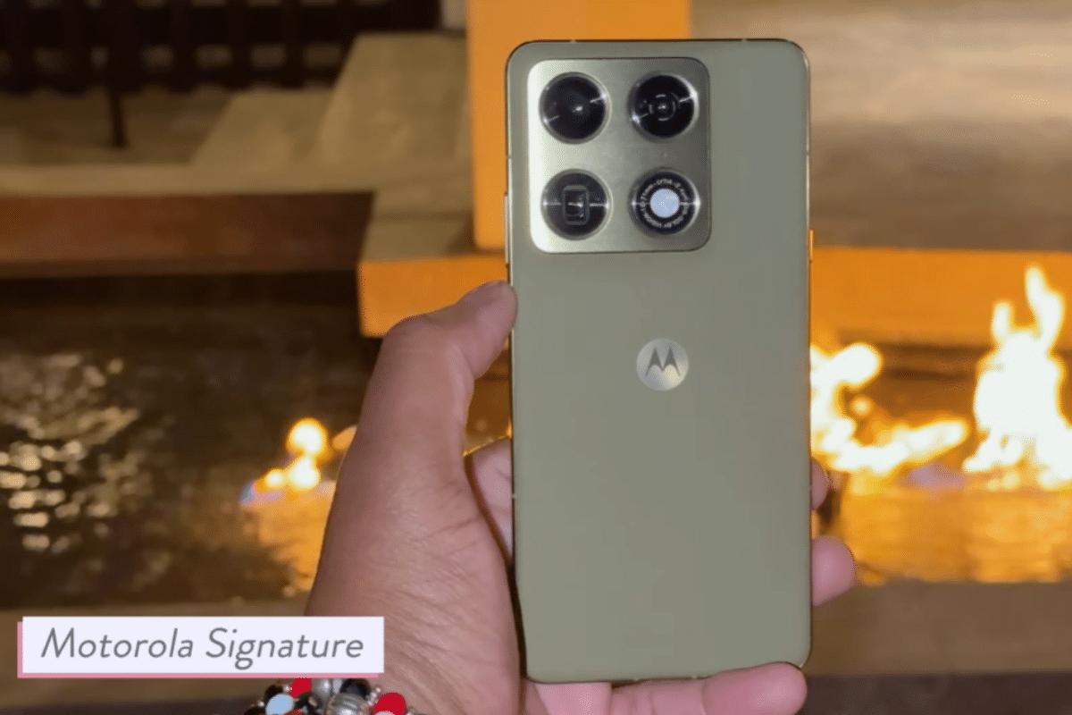 Mão segura Motorola Signature dourado com quatro câmeras, próximo a uma lareira acesa