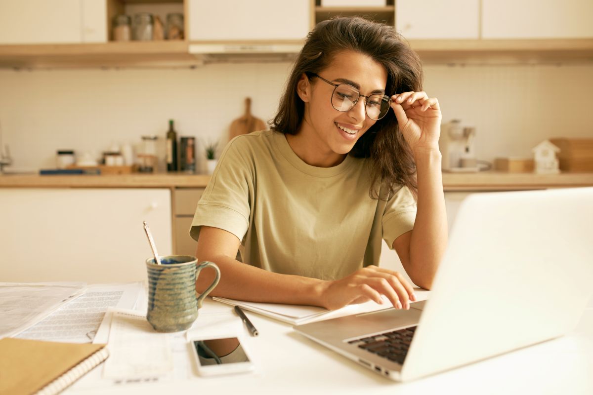 Jovem mulher sorrindo enquanto estuda em seu notebook, com livros e uma xícara de café sobre a mesa, simbolizando a preparação para o Enem.