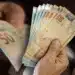 Mãos de idoso segurando e contando cédulas de reais em ambiente fechado.