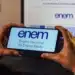 Pessoa segura celular exibindo tela do ENEM diante de notebook aberto na mesma página.