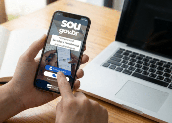 Pessoa utilizando celular com aplicativo SouGov aberto ao lado de notebook e caderno