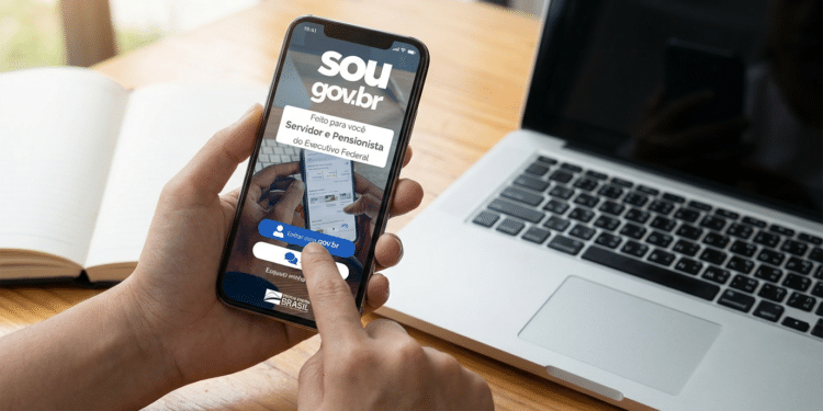 Pessoa utilizando celular com aplicativo SouGov aberto ao lado de notebook e caderno