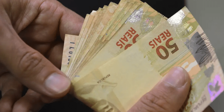 Mãos segurando cédulas de cinquenta reais em close representando o acréscimo de 30 por cento no salário dos trabalhadores com direito ao adicional de periculosidade a partir de abril de 2026