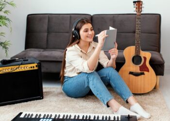 Jovem estudando inglês com música enquanto ouve fones de ouvido e faz anotações ao lado de um violão.