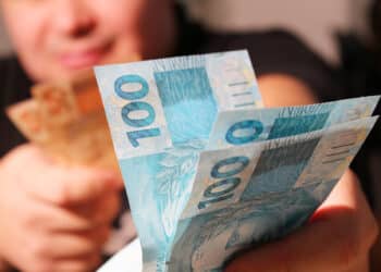 Mãos entregando notas de 100 reais destacadas em primeiro plano