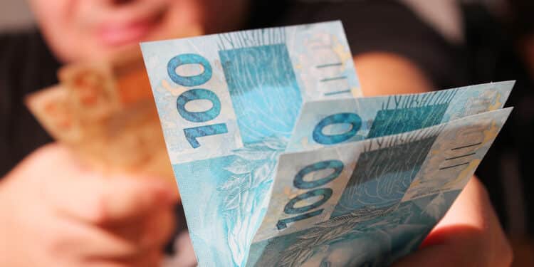 Mãos entregando notas de 100 reais destacadas em primeiro plano