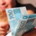 Mãos entregando notas de 100 reais destacadas em primeiro plano