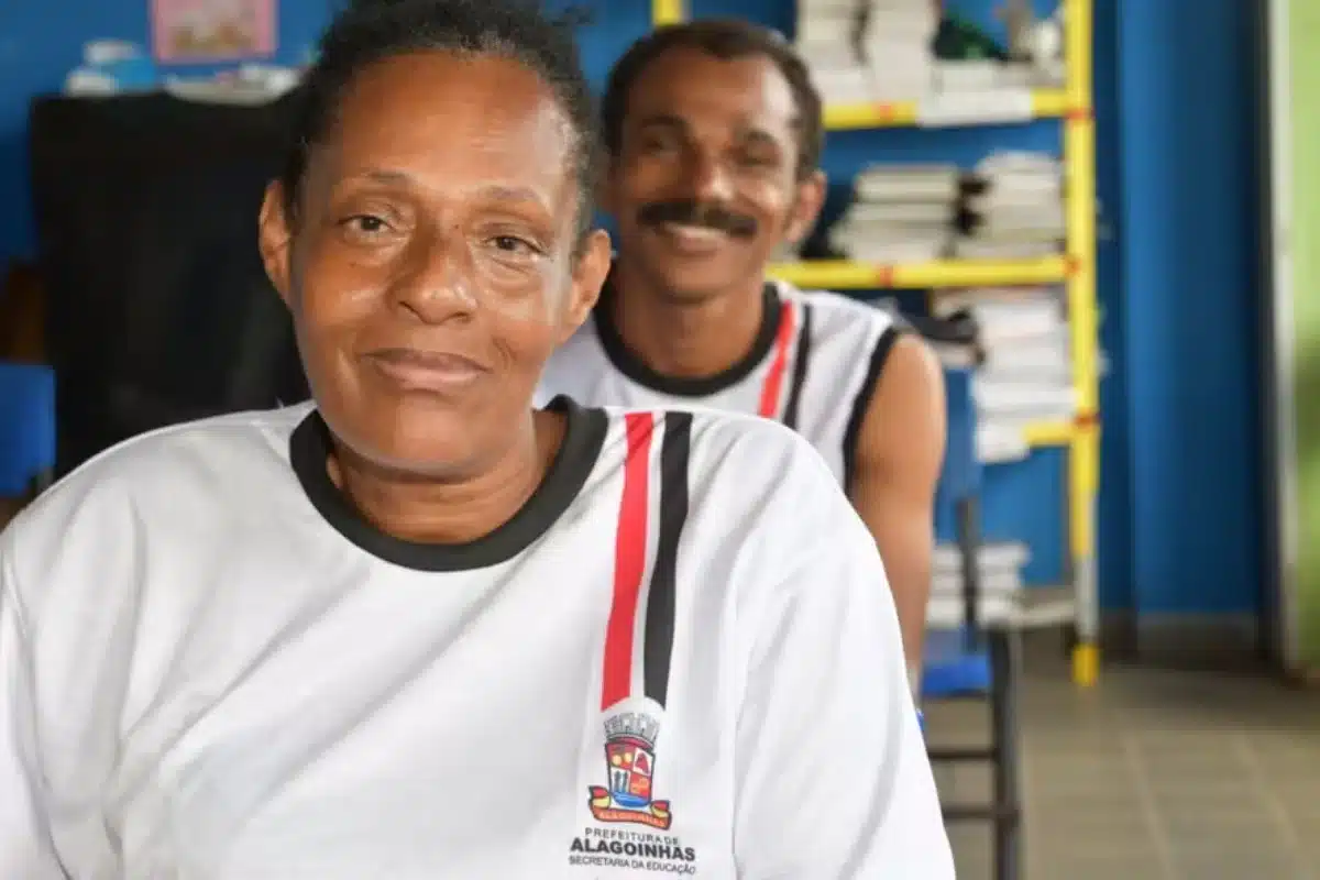 Dois adultos sorridentes usando camisetas brancas com o brasão da Prefeitura de Alagoinhas e identificação da Secretaria da Educação, sentados em ambiente escolar com estantes de livros ao fundo, representando estudantes da Educação de Jovens e Adultos beneficiados pelo programa Bolsa Presença
