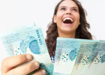 Mulher sorrindo segurando cédulas de 100 reais após anúncio de pagamento de R$ 950 pela Caixa para CPFs final 0 a 9