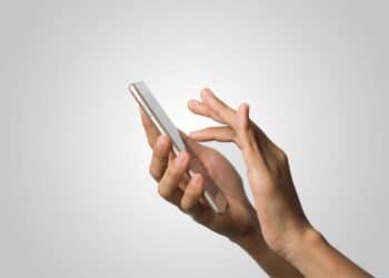 Close-up de mãos segurando um smartphone fino de cor dourada clara, com o dedo indicador tocando a tela, contra um fundo cinza neutro e minimalista.