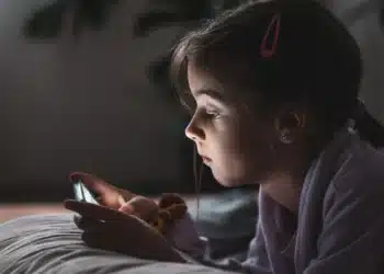 "Menina pequena usando um smartphone na cama, focada na tela, representando a questão do uso de dispositivos por crianças.